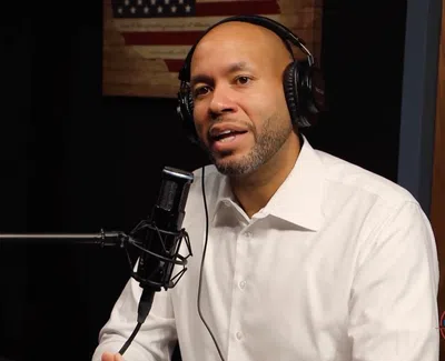 Man wearing headphones speaking into a microphone during a podcast recording with an American flag in the background.