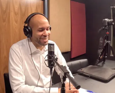 Man in white shirt with headphones speaking into a microphone in a podcast studio with acoustic panels.