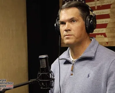 Man wearing headphones speaking into a microphone with a USA flag wall art in the background in a podcast studio