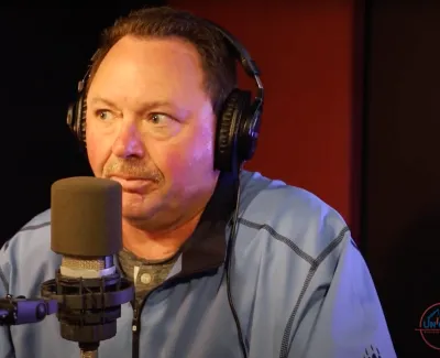 Man wearing headphones speaking into a microphone in a recording studio with podcast logos visible.