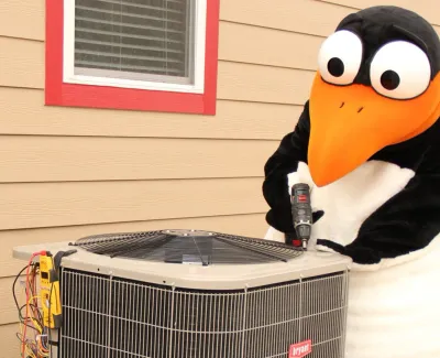 Person and penguin mascot using a drill to repair an air conditioning unit outside a beige house.