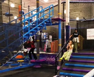 Children enjoying an indoor zipline ride with blue stairs and colorful padded steps in a play area.