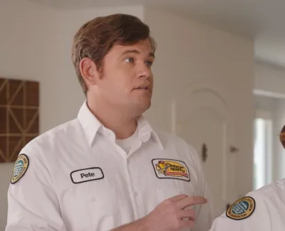 Two cleaning service professionals in white uniforms with logos and name tags inside a residential home.