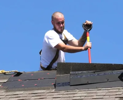 Construction worker using roofing tool to install new shingles on rooftop under clear blue sky