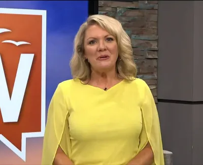 Smiling blonde woman in yellow dress standing in TV studio with a colorful backdrop and stone wall