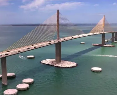 Aerial view of a cable-stayed bridge over calm blue water with vehicles and a boat passing beneath overclear skies.