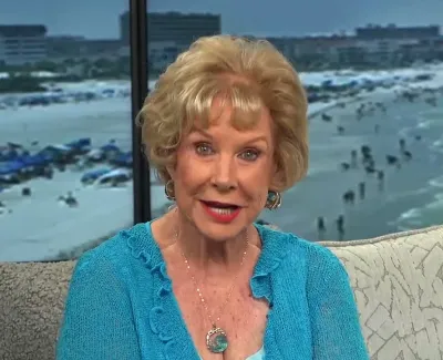Senior woman with short blonde hair wearing a turquoise sweater speaking indoors with a beach scene visible through windows.