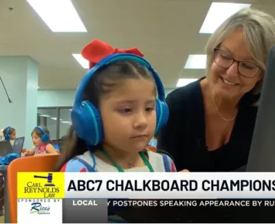 Young girl wearing headphones using a laptop with teacher assisting her in a classroom setting