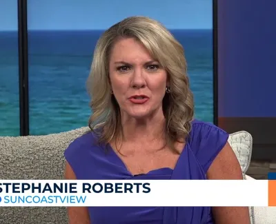 News anchor Stephanie Roberts in a blue dress delivering a report on Suncoast View with ocean backdrop.