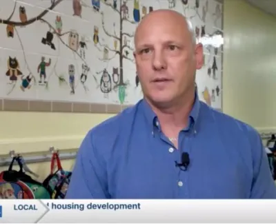 Man in blue shirt speaks in a school hallway with children's backpacks and mural of owls behind him.
