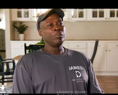 Man wearing a James Dia Services Do It All shirt sitting in a kitchen with white cabinets and chairs.
