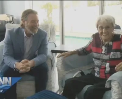Man in blue suit and elderly woman in patterned shirt smiling while sitting in living room chairs by window.