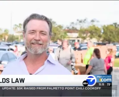 Reporter Carl Reynolds smiling during a live news segment with a crowd and cars in the background.