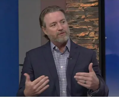 Man in a suit speaking and gesturing during a television interview on a local news set.