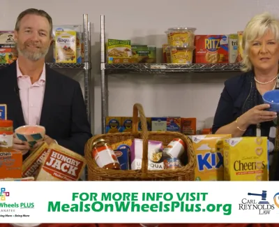 Two people holding bowls surrounded by various packaged food items promoting Meals on Wheels Plus program.