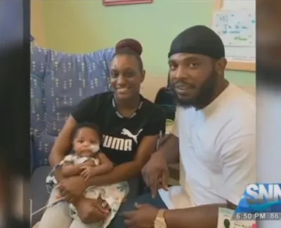 Happy family of three sitting together indoors, smiling at the camera, with baby in mother's arms.