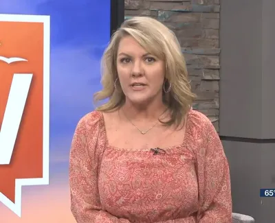 Blonde female news anchor in patterned pink blouse presenting in front of a blue and orange backdrop with temperature and time shown.