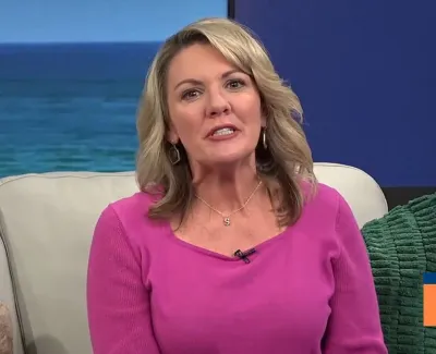 Blonde woman in a pink sweater speaking on a TV show set with ocean background and green pillow.