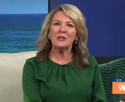 Woman with blonde hair in green blouse speaking on a TV set with ocean background and Suncoast View logo.