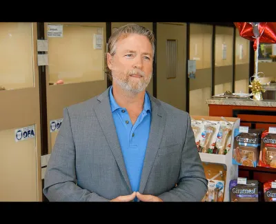 Man in gray suit and blue shirt stands in pet store near shelves with Gimme's pet treats and red star balloon.