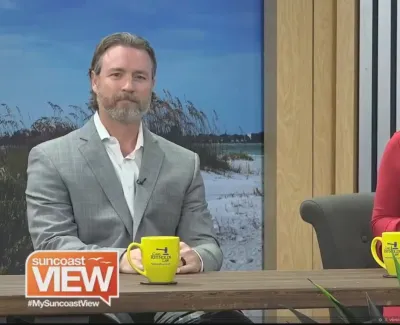 Two Suncoast View hosts seated at a desk with yellow mugs and a beach backdrop in a TV studio