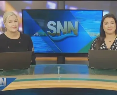 Two female news anchors seated at a news desk with laptops and SNN logo on a screen behind them.