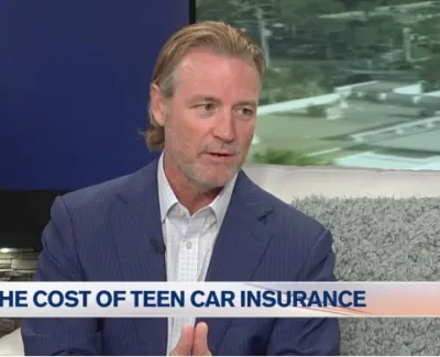 Man in blue suit discussing the cost of teen car insurance on a TV set with a city view background.