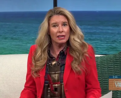 Woman with long blonde hair in red blazer sits on white couch holding a vintage trophy with ocean backdrop