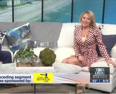 Two hosts sitting on a white couch in a studio with ocean view windows during a local news segment.