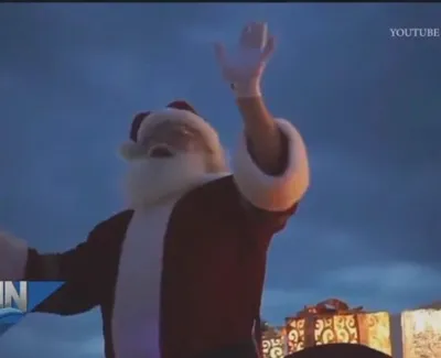 Santa Claus waving joyfully with illuminated holiday decorations in the background during early morning.