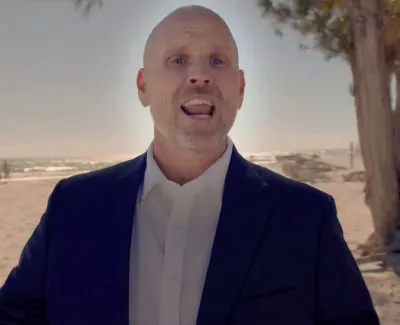 Man in a suit speaking on a sunny beach with palm trees and ocean in the background.