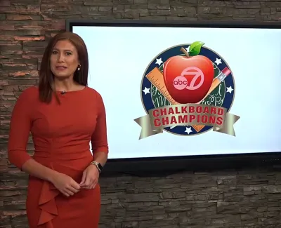 Two newscasters standing beside a screen displaying ABC 7 Chalkboard Champions logo with an apple and pencil graphic.