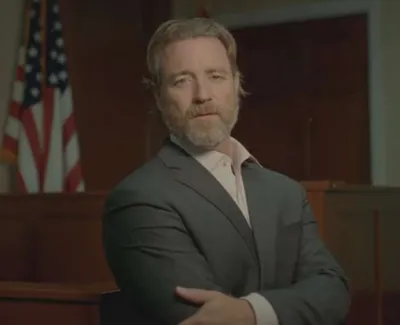 Man with beard and suit standing confidently in courtroom with American flag in background