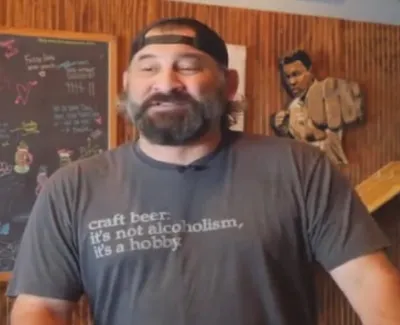 Man with beard and backward cap wearing a craft beer hobby t-shirt in a bar with wooden walls and chalkboard.