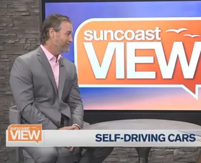 Two TV hosts discuss self-driving cars on Suncoast View with a studio backdrop and temperature display.