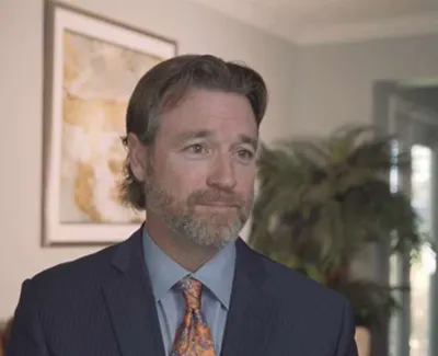 Man in a suit and patterned tie standing in a modern room with artwork and plants in the background
