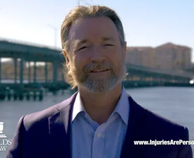 Man in a suit smiling near a waterfront with a bridge and buildings in the background, Carl Reynolds Law logo visible.