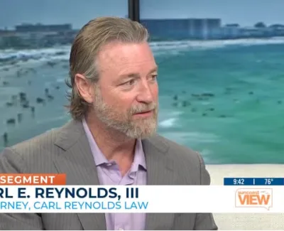 Man in a suit appearing on a daytime talk show with ocean and beach background, sponsored segment for Carl Reynolds Law