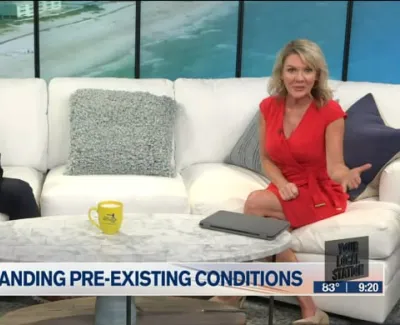 Two TV hosts seated on a white couch with a beach backdrop discussing pre-existing medical conditions