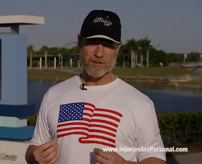 Man wearing American flag shirt and Carl Reynolds Law cap stands outdoors near American Youth Cup sign at Nathan Benderson Park.