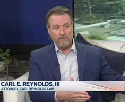 Attorney Carl E. Reynolds III discussing legal matters on a TV news set with plants and a lamp in the background.