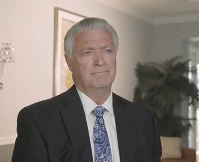 Older man in a dark suit and paisley tie sitting in a modern, softly lit living room with neutral decor.
