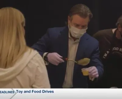 Volunteers wearing masks serve food during a toy and food drive event under health safety measures.