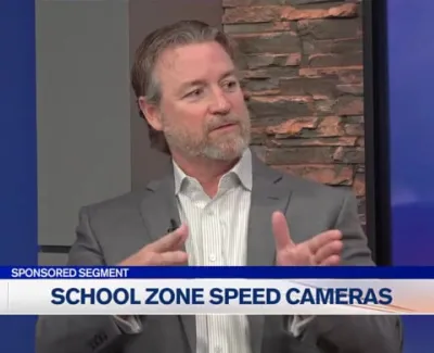 Man in gray suit discussing school zone speed cameras on local news broadcast segment.