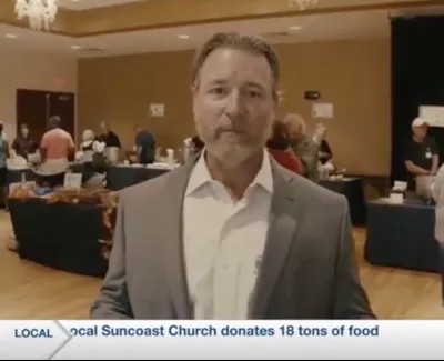 Man in a suit speaks at an indoor event where a church donates 18 tons of food with people and tables in the background