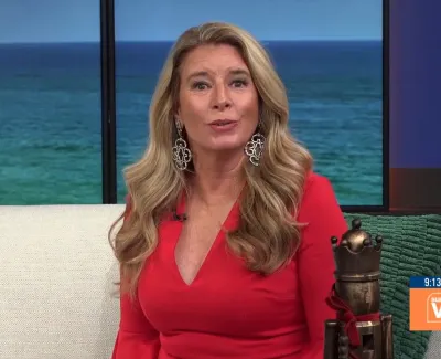 Female TV host with long blonde hair in red dress sitting on couch with ocean backdrop on Suncoast View show.
