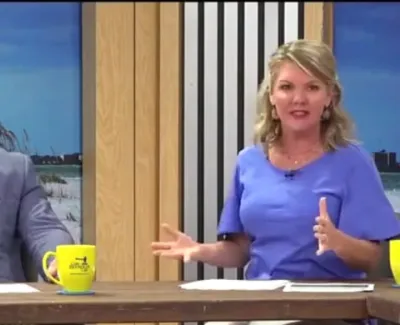 Man and woman hosting a talk show at a wooden desk with yellow mugs and a beach backdrop.