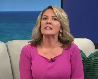 Blonde woman in pink sweater speaking on a beige couch with green pillow and ocean background.