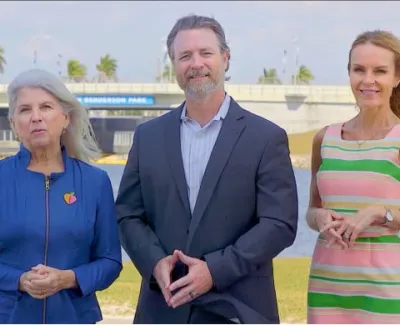 Three people standing outdoors by a waterfront with a bridge and palm trees in the background.