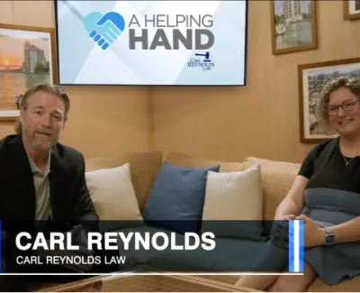 Two people sitting in a cozy room with A Helping Hand logo on screen behind them and Carl Reynolds Law caption.